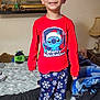 child, boy, smiling, pajamas, bed, blanket, lamp, painting, room, furniture, toy, red_clothing, blue_pants, snowflake_pattern, socks, smile, standing, indoors, person, home