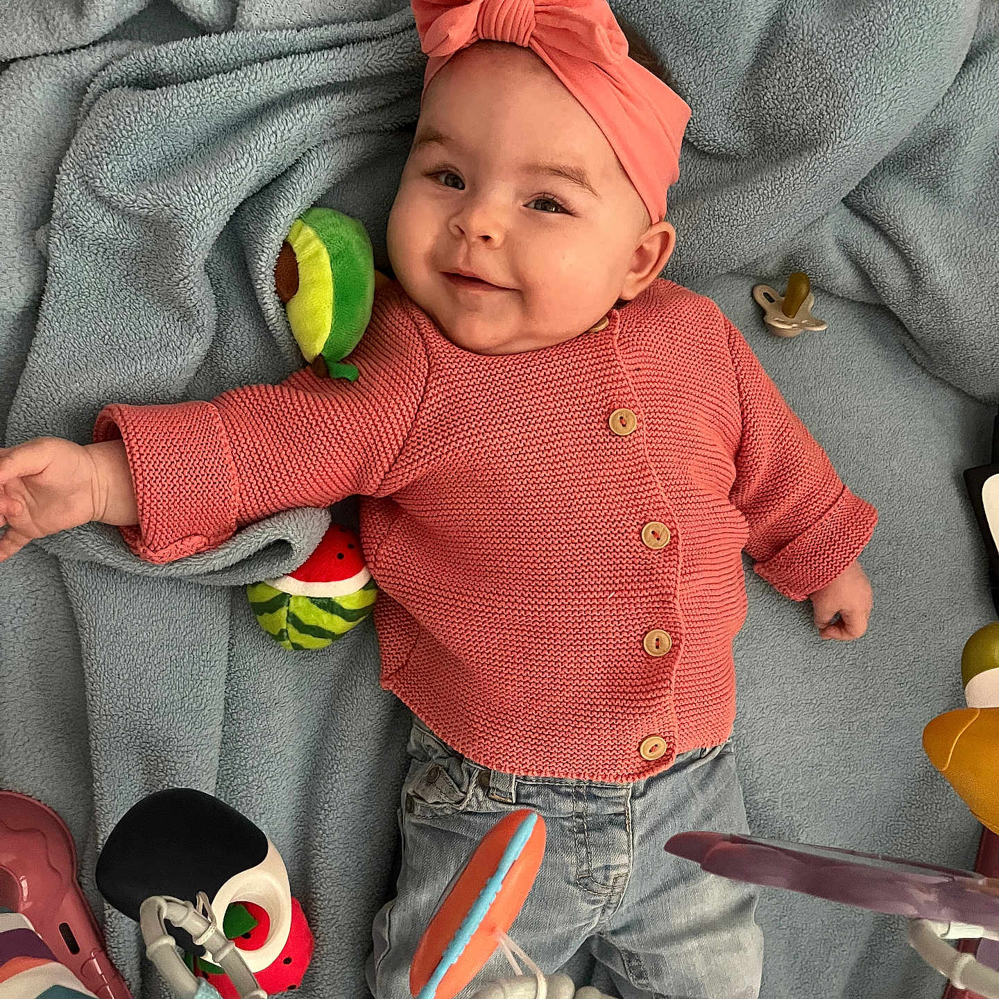 Tiya participe au concours pour gagner de l'argent avec cette photo : avocado_toy, baby, blue_blanket, buttoned_sweater, child, coral_sweater, cute, happy, headband, indoors, infant, jeans, pacifier, playful, plush_toys, portrait, smiling, soft_toy, toy_bar, watermelon_toy