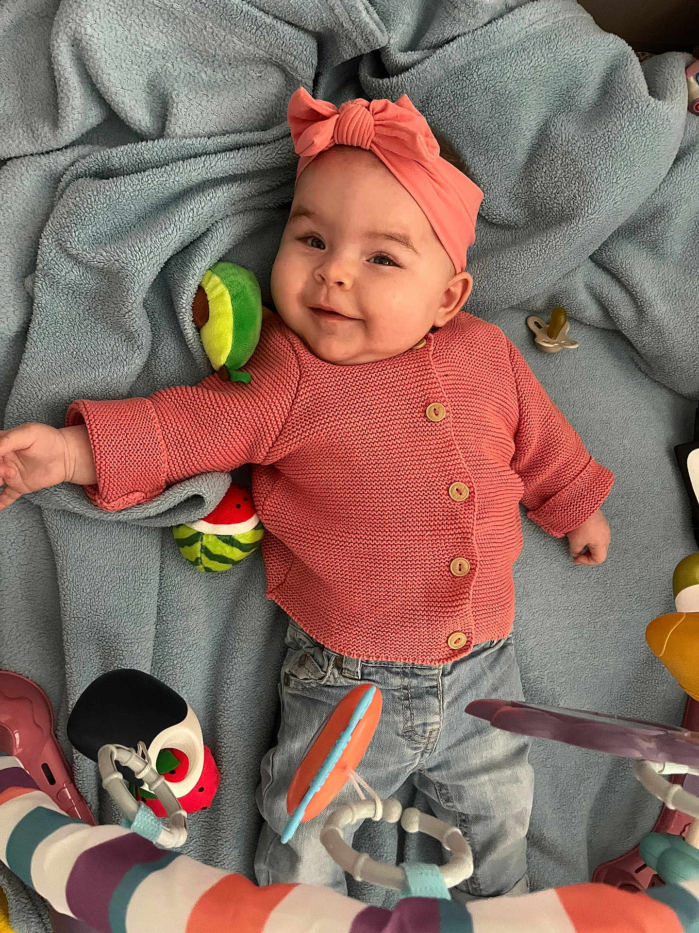 Tiya participe au concours pour gagner de l'argent avec cette photo : baby, smiling, coral_sweater, jeans, blue_blanket, plush_toys, avocado_toy, watermelon_toy, pacifier, soft_toy, infant, cute, child, playful, indoors, portrait, happy, headband, buttoned_sweater, toy_bar