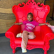 Lyna a rejoint le concours — aidez-le/la à gagner de superbes lots ! braided_hair, casual_clothing, child, comfortable, decor, fashion, floor_tiles, furniture, indoor, pink_pants, portrait, red_chair, relaxed_pose, seated, smiling, sneakers, throne, tshirt, wooden_wall, young_person