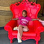 braided_hair, casual_clothing, child, comfortable, decor, fashion, floor_tiles, furniture, indoor, pink_pants, portrait, red_chair, relaxed_pose, seated, smiling, sneakers, throne, tshirt, wooden_wall, young_person