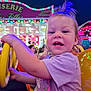 Lorena a rejoint le concours — aidez-le/la à gagner de superbes lots ! toddler, child, amusement_ride, steering_wheel, carnival, fairground, lights, purple_shirt, unicorn_graphic, happy, smiling, hair_tuft, people, night, fun, colorful, yellow, orange, playful, crowd