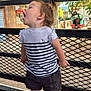 toddler, child, striped_shirt, shorts, sandals, metal_fence, outdoor, tree, toy_train, playground, sunlight, hair_tie, expression, side_profile, candid, nature, summer, young_child, shoes, fence