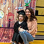 Charlee joined the competition — help win amazing prizes! child, woman, graduation_gown, cap, indoor, wooden_floor, colorful_backdrop, smiling, casual_clothing, jeans, sneakers, celebration, school_theme, portrait, people, happy, curly_hair, kneeling, holding, young