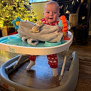 Marylynn is registered to the contest to win money with this photo: baby, walker, toy, blanket, christmas_tree, lights, pajamas, floor, indoor, smile, face, person, furniture, window, curtain, chair, wooden_floor, holiday, festive, home