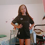 Kayliah participe au concours pour gagner de l'argent avec cette photo : bed, bedroom, black_shirt, casual, child, confident, furniture, girl, hands_on_hips, home, indoor, long_hair, pillow, portrait, posing, relaxed, stroller, toys, wall_decor, young_person