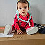 toddler, child, baby, red_clothing, onesie, headband, cardboard_box, indoor, curious, sitting, footwear, pink_shoes, toy_accessory, playful, young_child, expression, wall_background, casual, home, person