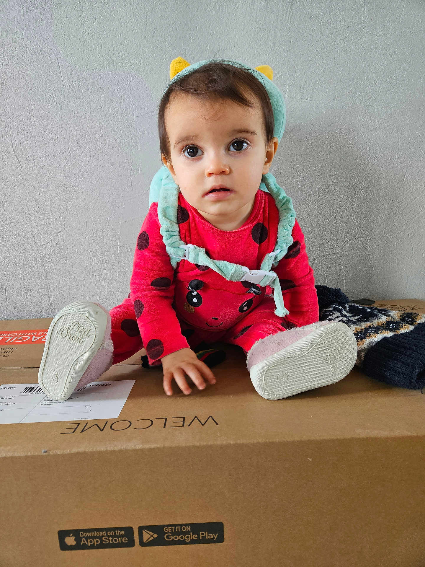 Rose participe au concours pour gagner de l'argent avec cette photo : toddler, child, baby, red_clothing, onesie, headband, cardboard_box, indoor, curious, sitting, footwear, pink_shoes, toy_accessory, playful, young_child, expression, wall_background, casual, home, person