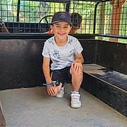 Hugo participe au concours pour gagner de l'argent avec cette photo : child, boy, smiling, cap, white_tshirt, black_shorts, sneakers, toy, vehicle, metal_floor, mesh_barrier, wooden_structure, outdoor, greenery, daylight, casual_clothing, happy, portrait, playful, seated
