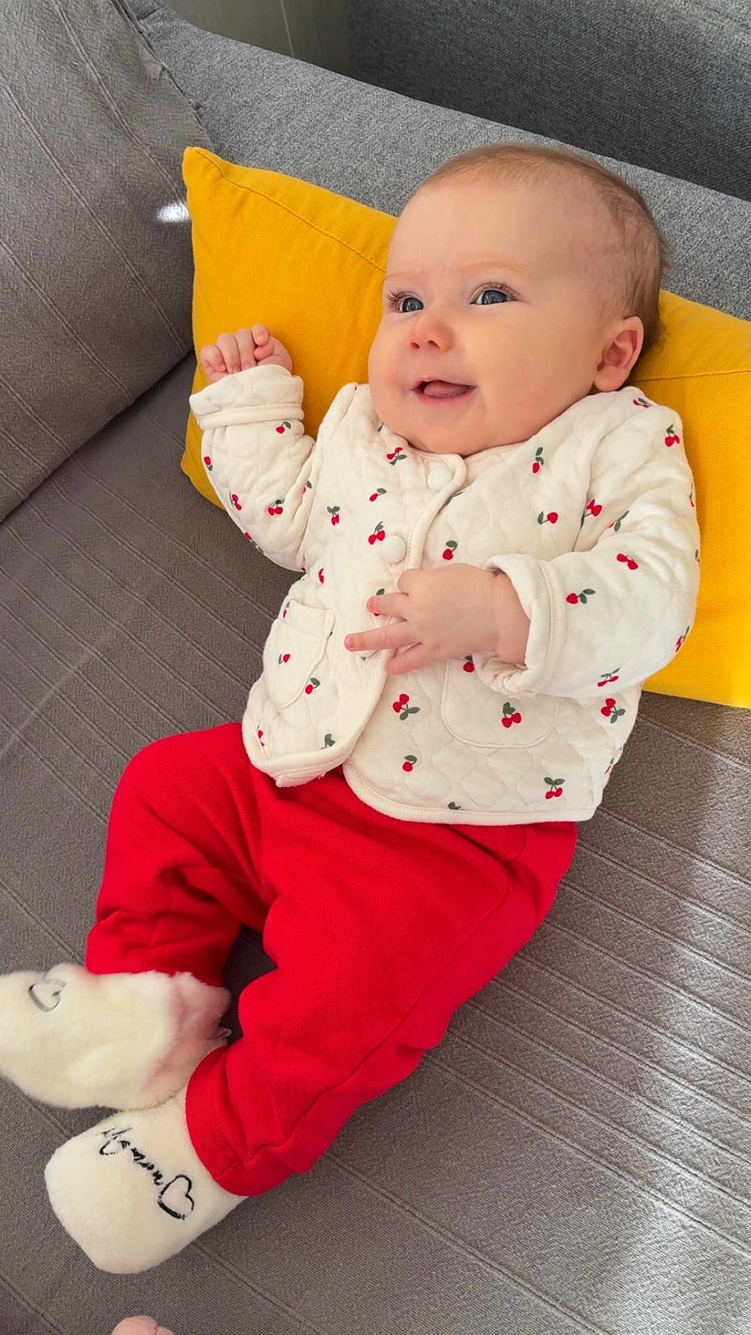Lila participe au concours pour gagner de l'argent avec cette photo : baby, infant, smiling, yellow_pillow, grey_couch, red_pants, white_jacket, cherry_pattern, socks, slippers, hands, eyes, portrait, cozy, home_interior, fabric, cushion, happy, seated, cute