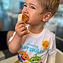 child, toddler, boy, eating, toast, food, shirt, baby_shark, smiling, cute, indoors, short_hair, hand, face, person, clothing, sitting, bright, colorful, happy