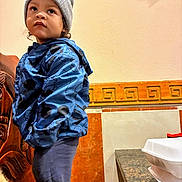 Marcus is registered to the contest to win money with this photo: child, toddler, beanie, gray_hat, blue_jacket, patterned_jacket, standing, indoor, wall, decorative_tile, countertop, takeout_container, table, looking_up, thoughtful_expression, portrait, pants, wooden_furniture, curly_hair, casual_clothing