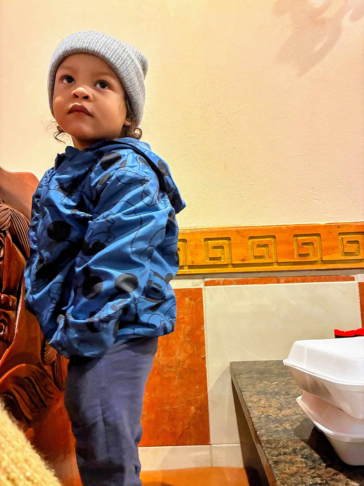 Marcus is registered to the contest to win money with this photo: child, toddler, beanie, gray_hat, blue_jacket, patterned_jacket, standing, indoor, wall, decorative_tile, countertop, takeout_container, table, looking_up, thoughtful_expression, portrait, pants, wooden_furniture, curly_hair, casual_clothing