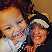 Marcus is registered to the contest to win money with this photo: child, adult, smiling, selfie, curly_hair, toothy_grin, cap, hat, indoor, couch, pillow, close_up, portrait, hand, family, jacket, t_shirt, zipper, happy, eyes