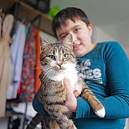 Alpha participe au concours pour gagner de l'argent avec cette photo : blurred_background, casual_wear, cat, child, clothing, colorful, cute, domestic_animal, furniture, green_eyes, happy, holding, indoor, person, pet, portrait, shelf, smiling, tabby_cat, white_paws