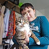 blurred_background, casual_wear, cat, child, clothing, colorful, cute, domestic_animal, furniture, green_eyes, happy, holding, indoor, person, pet, portrait, shelf, smiling, tabby_cat, white_paws