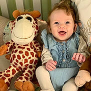 Eve joined the competition — help win amazing prizes! baby, child, smiling, stuffed_animal, giraffe, plush_toy, blue_dress, white_tights, curly_hair, happy, indoor, seated, cushion, soft_toys, playroom, portrait, cute, infant, cozy, home