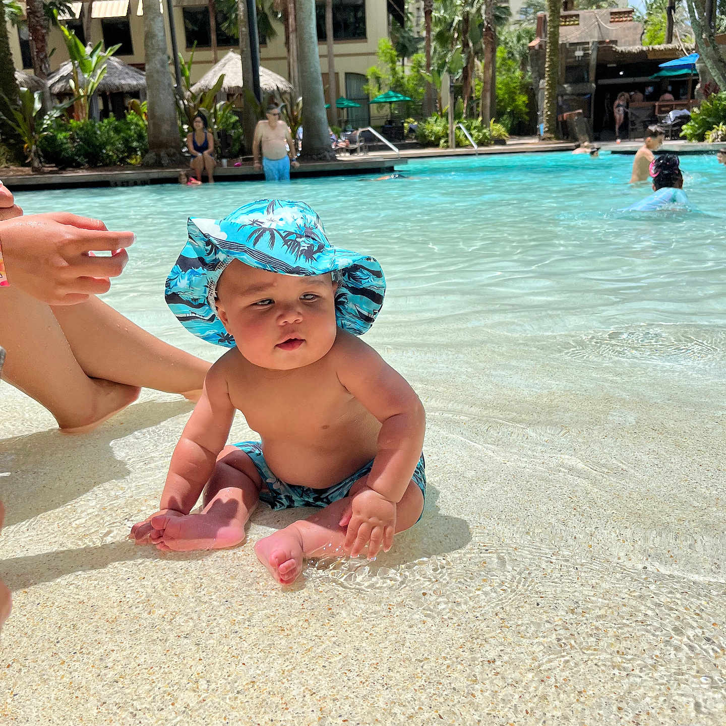 Zion joined the competition — help win amazing prizes! baby, child, infant, swimming_pool, resort, outdoor, tropical, palm_trees, sunhat, swimwear, water, sitting, smiling, portrait, family, vacation, sunlight, shallow_water, poolside, people