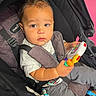 Zion joined the competition — help win amazing prizes! baby, child, infant, stroller, seated, portrait, face, wide_eyed, serious_expression, short_hair, toy, colorful_toy, harness, seatbelt, outdoor, pink_background, fabric, clothing, photography, close_up