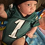baby, infant, child, sports_jersey, green_clothing, hat, blanket, colorful, sitting, indoors, portrait, cute, young_child, soft_focus, face, human, one, number, hand, leg