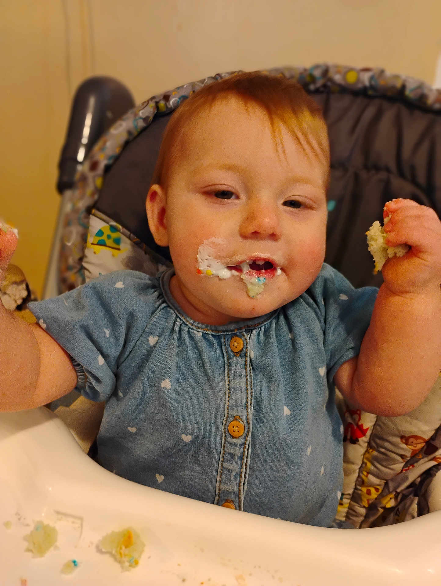 Blanche is registered to the contest to win money with this photo: baby, child, high_chair, cake, frosting, crumbs, blue_dress, heart_pattern, happy, messy, food, eating, cheeks, hands, indoor, person, cute, face, infant, smiling