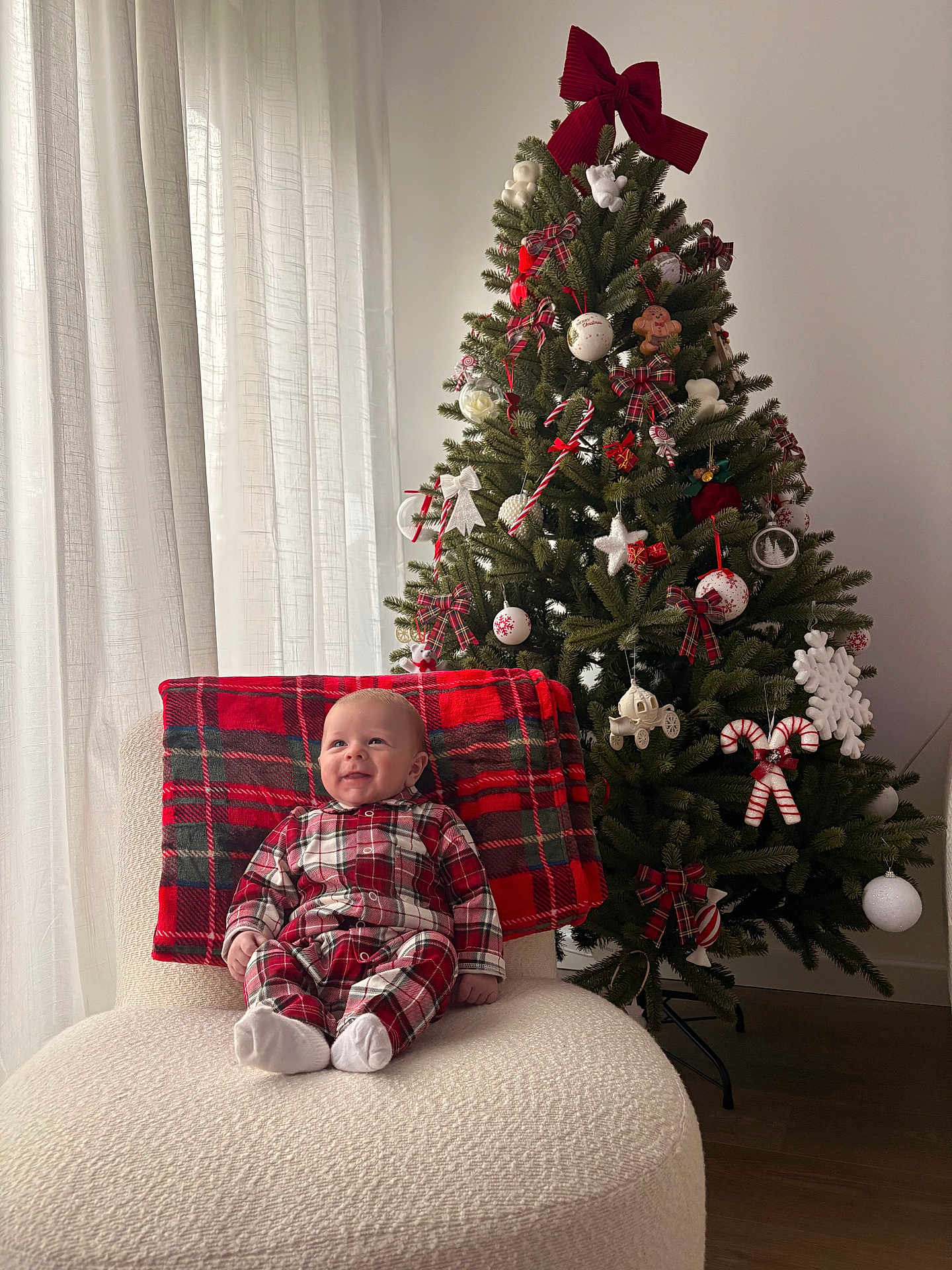 Raphaël a rejoint le concours — aidez-le/la à gagner de superbes lots ! baby, child, plaid_pajamas, chair, blanket, christmas_tree, ornaments, candy_cane, bow, holiday, smile, happy, indoor, curtains, decorations, festive, person, sitting, cozy, wood_floor