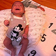 Milo joined the competition — help win amazing prizes! baby, infant, smile, laughing, mickey_mouse, romper, pillow, blanket, numbers, floor, wooden_floor, clothing, cute, happy, child, person, indoor, portrait, skin, head