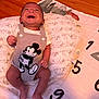 baby, infant, smile, laughing, mickey_mouse, romper, pillow, blanket, numbers, floor, wooden_floor, clothing, cute, happy, child, person, indoor, portrait, skin, head