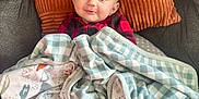 Logan is registered to the contest to win money with this photo: baby, blanket, cushion, red_plaid, orange_pillow, armchair, cute, smile, cozy, indoor, person, child, footwear, soft_toys, patterned_blanket, head, face, comfort, happy, toddler