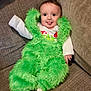 Logan joined the competition — help win amazing prizes! baby, child, green_furry_outfit, tongue_out, couch, smiling, happy, indoor, infant, person, cute, plush, clothing, sitting, funny_face, young_child, cozy, home, cushion, soft