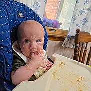 Colton joined the competition — help win amazing prizes! baby, high_chair, messy, food, bib, hand, face, indoor, curious, kitchen, curtains, window, wooden_chair, table, feeding, child, mess, infant, eating, smudged