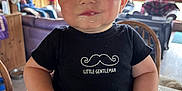 Ivan is registered to the contest to win money with this photo: baby, child, high_chair, black_shirt, mustache_logo, drool, indoor, wooden_interior, natural_light, smile, cute, face, person, furniture, table, window, cozy, home, blue_eyes, expression