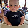 baby, child, high_chair, black_shirt, mustache_logo, drool, indoor, wooden_interior, natural_light, smile, cute, face, person, furniture, table, window, cozy, home, blue_eyes, expression