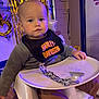 Ivan is registered to the contest to win money with this photo: baby, child, high_chair, bib, harley_davidson, face, indoor, balloon, decorations, purple_light, table, clothing, pacifier, wood_floor, expressionless, person, seat, party, toddler, brown_pants