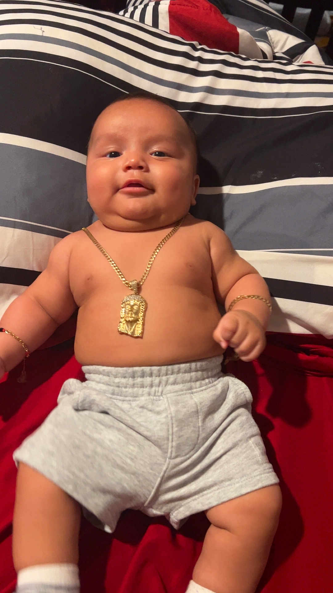 Krisniel is registered to the contest to win money with this photo: baby, infant, child, gold_chain, bracelet, shorts, socks, bed, pillow, striped_pillow, red_blanket, skin, face, chubby, smiling, lying_down, indoors, cute, jewelry, comfort