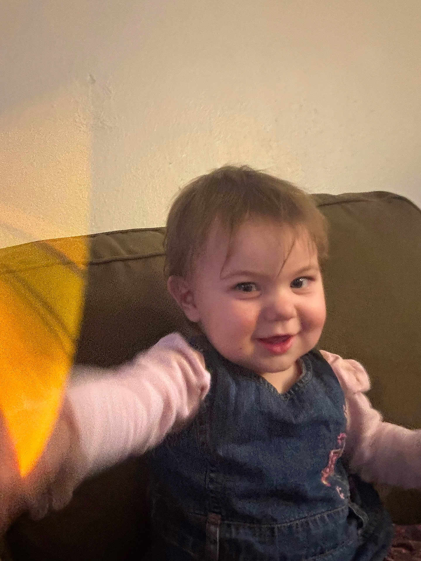 Aryella joined the competition — help win amazing prizes! toddler, child, smile, face, couch, indoor, clothing, denim, pink_sleeves, hair, happy, young_child, person, portrait, seated, wall, warm_light, reaching_hand, casual, home