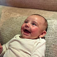 Dani participe au concours pour gagner de l'argent avec cette photo : baby, infant, smile, laughing, onesie, couch, cushion, face, eyes, cheeks, hand, ear, portrait, closeup, indoor, fabric, happy, expression, skin, blurry_background