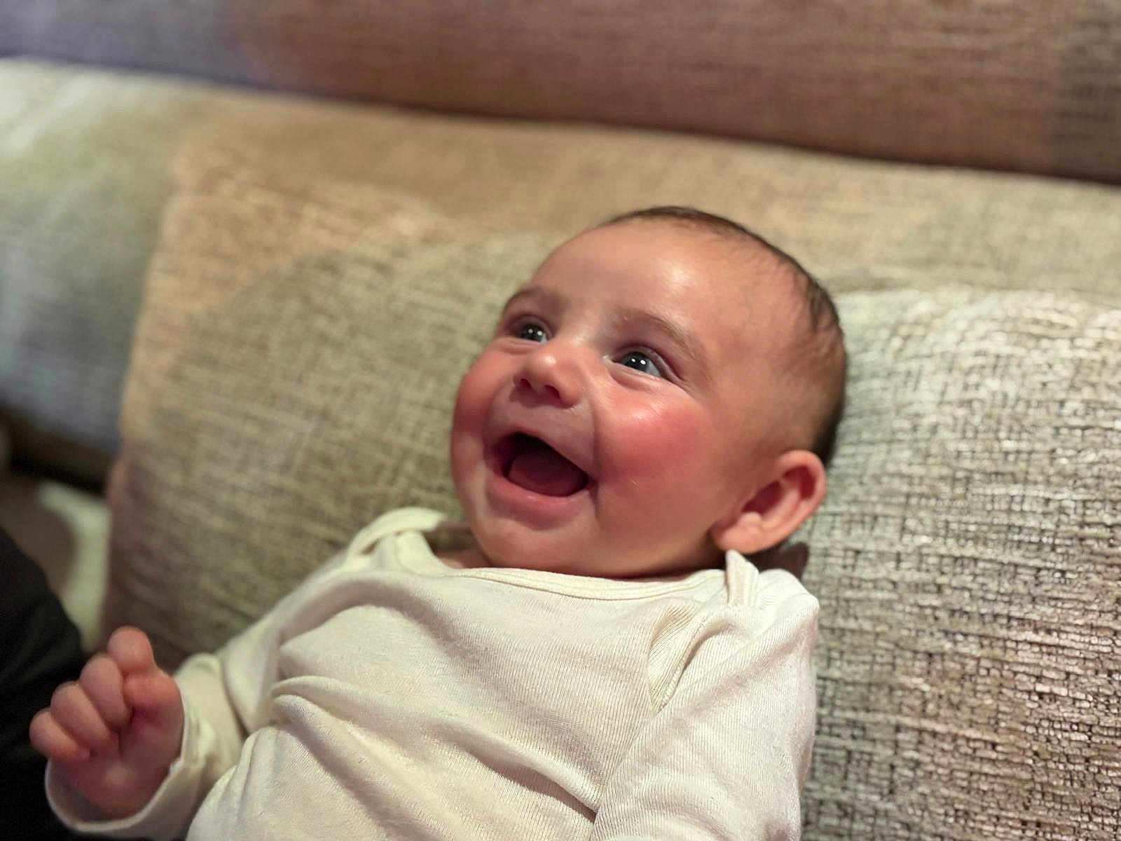 Dani participe au concours pour gagner de l'argent avec cette photo : baby, infant, smile, laughing, onesie, couch, cushion, face, eyes, cheeks, hand, ear, portrait, closeup, indoor, fabric, happy, expression, skin, blurry_background