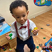 Kaizir is registered to the contest to win money with this photo: toddler, child, boy, indoors, playroom, toys, wooden_floor, window, bow_tie, suspenders, shirt, pants, sneakers, plastic_toys, activity_board, smiling, standing, curly_hair, person, play