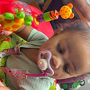 Nylayah is registered to the contest to win money with this photo: toddler, pacifier, toy, baby_seat, child, indoor, play, colorful, person, arm, floor, wooden_floor, clothing, green, orange, purple, furniture, blurred_background, home, sitting