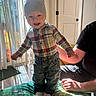 baby, child, smiling, standing, indoor, portrait, plaid_shirt, beanie, pants, socks, quilt, bed, adult, parent, sunlight, window, blinds, home, wood_floor, family