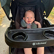 Cole is registered to the contest to win money with this photo: baby, child, infant, stroller, smiling, outdoor, park, path, seated, portrait, face, eyes, striped_shirt, carriage, canopy, safety_harness, cup_holder, black_stroller, happy, daylight
