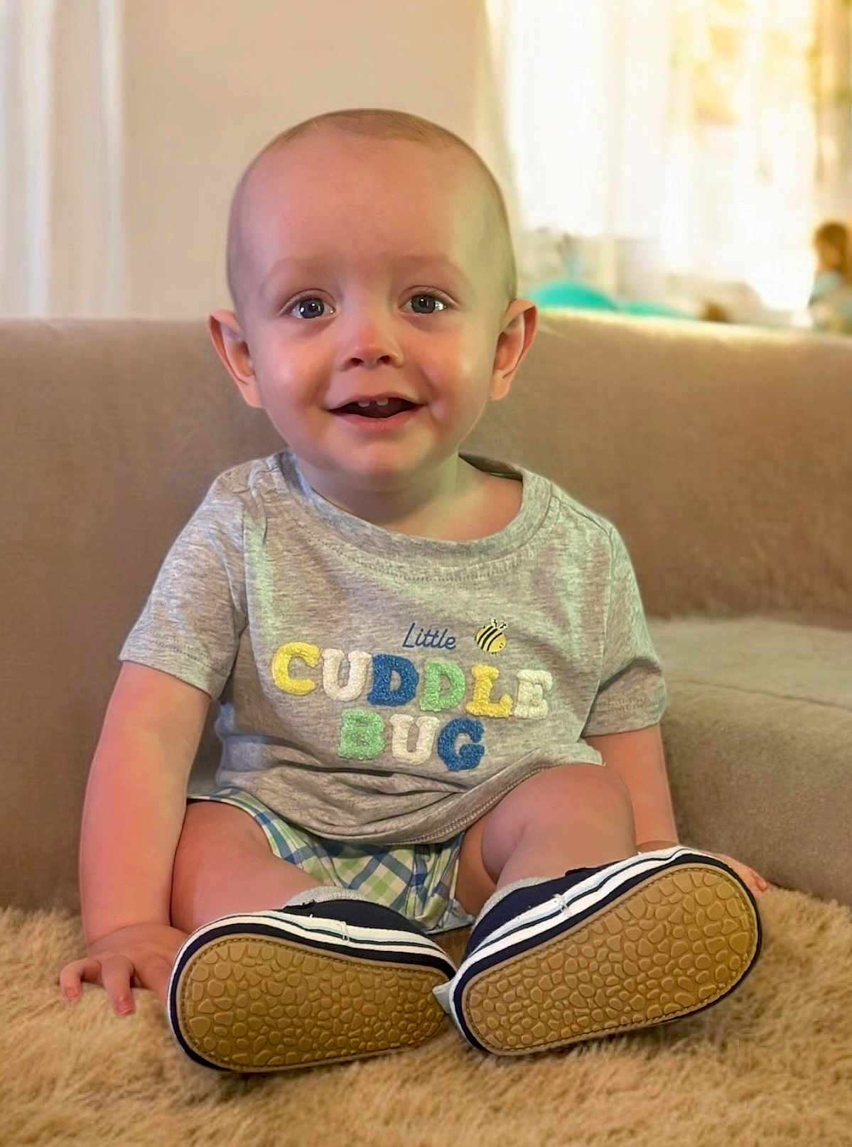 Rowan joined the competition — help win amazing prizes! toddler, child, smiling, sitting, indoor, shirt, text_on_clothing, shoes, plaid_shorts, rug, furniture, couch, happy, face, person, cute, baby, young_child, casual_wear, bright