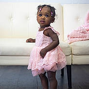 Lana is registered to the contest to win money with this photo: toddler, child, dress, pink_dress, sandals, floor, wooden_floor, couch, furniture, blanket, pink_blanket, indoor, person, cute, standing, beads, hair_accessories, wall, white_wall, curious