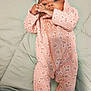 baby, infant, child, onesie, pink_clothing, headband, bow, face, hands, lying_down, quilted_blanket, cute, soft_fabric, patterned_clothing, footed_pajamas, adorable, indoors, portrait, young_child, playful
