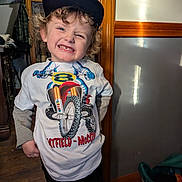 Jacobi joined the competition — help win amazing prizes! child, smiling, curly_hair, cap, tshirt, indoor, wooden_floor, wooden_panel, cabinet, toy, person, happy, face, casual_clothing, room, light, curtain, furniture, playful, portrait