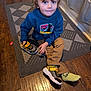 child, curly_hair, blue_hoodie, sneakers, yellow_shoes, green_socks, rug, wooden_floor, flooring, indoor, home, kitchen, patterned_rug, sitting, cross_legged, young_child, casual_clothing, person, footwear, candid