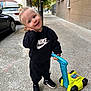 toddler, child, smiling, outdoor, sidewalk, toy, lawn_mower, sneakers, black_clothing, hoodie, pants, blonde_hair, blue_eyes, urban, trees, cars, building, playful, cute, person