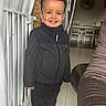 child, boy, toddler, smiling, portrait, standing, indoors, home, winter_clothing, jacket, dark_clothing, curly_hair, happy, face, radiator, curtain, sofa, dining_area, chairs, floor