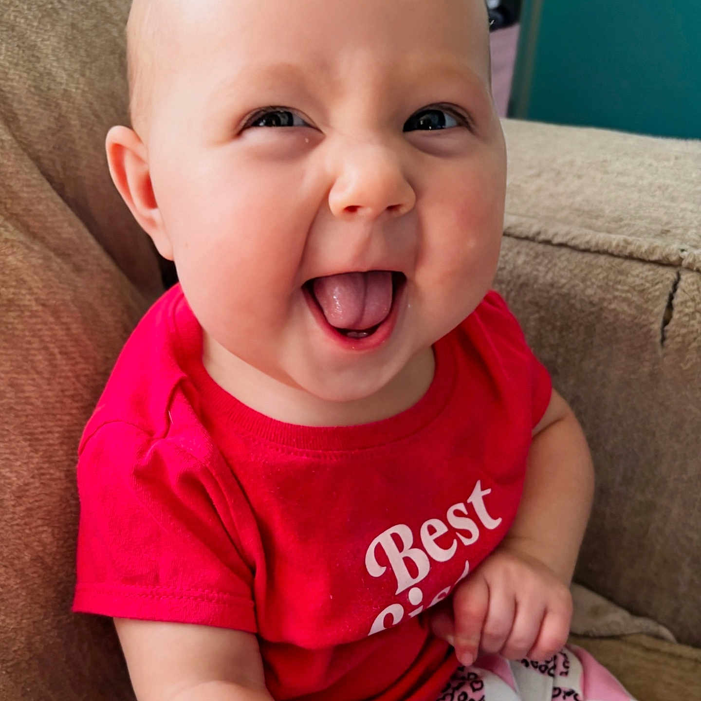 Vhonda is registered to the contest to win money with this photo: baby, child, smiling, tongue_out, red_shirt, sitting, couch, indoor, face, happy, person, patterned_pants, furniture, head, skin, cute, young_child, portrait, clothing, infant